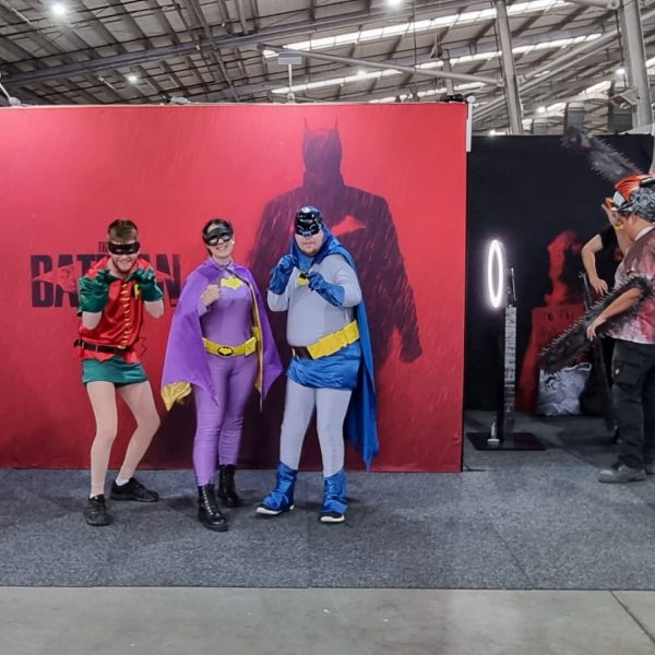 three people dressed in costumes in front of a batman media wall at a fan event