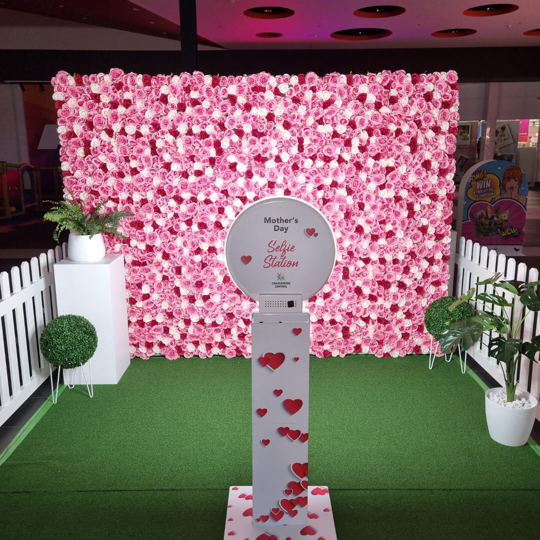 flower wall set up inside a white picket fence with other decorations and a photo booth for a mothers day event at a shopping centre